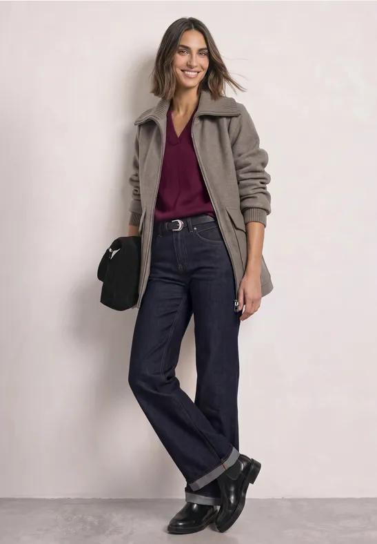 Frau in grauer Jacke, Bordeaux-Shirt und weiten Jeans, schwarze Stiefel und Tasche, lächelnd stehend.