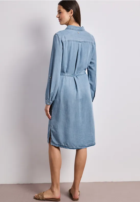 Vrouw van achteren in een knielange spijkerjurk met lange mouwen, blootsvoets.