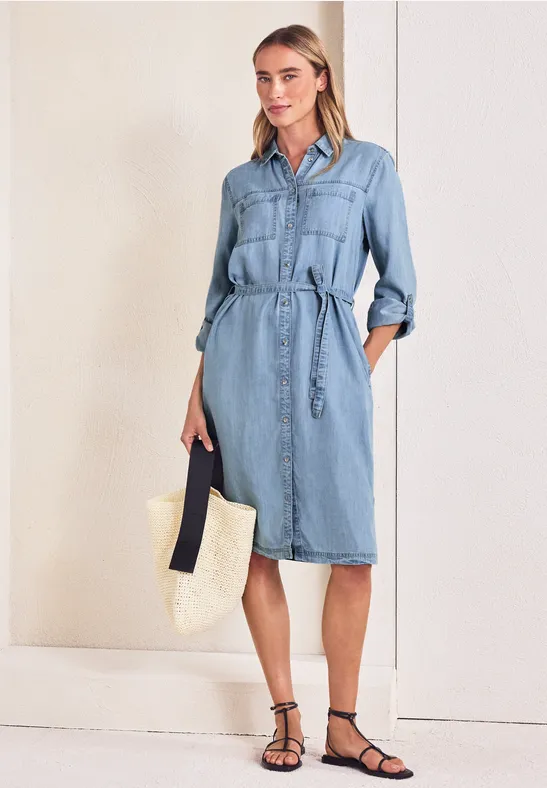 Vrouw in lichtblauwe denim jurk met ceintuur, draagt een stro-tas en zwarte sandalen.