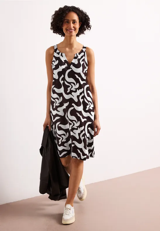 Vrouw met kort krullend haar draagt een mouwloze, knielange jurk met zwart-wit patroon en witte sneakers.