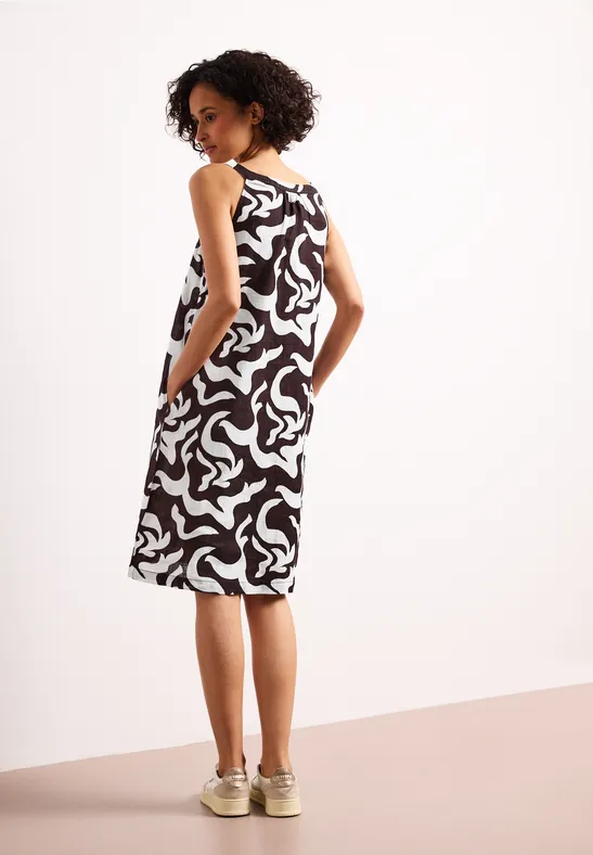 Vrouw van achteren in een zwart-witte, mouwloze midijurk met abstract patroon, zakken en beige sneakers.