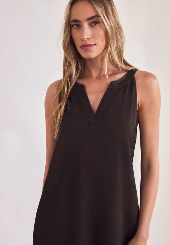 Vrouw met lang blond haar in een mouwloze zwarte jurk met V-hals tegen een neutrale achtergrond.