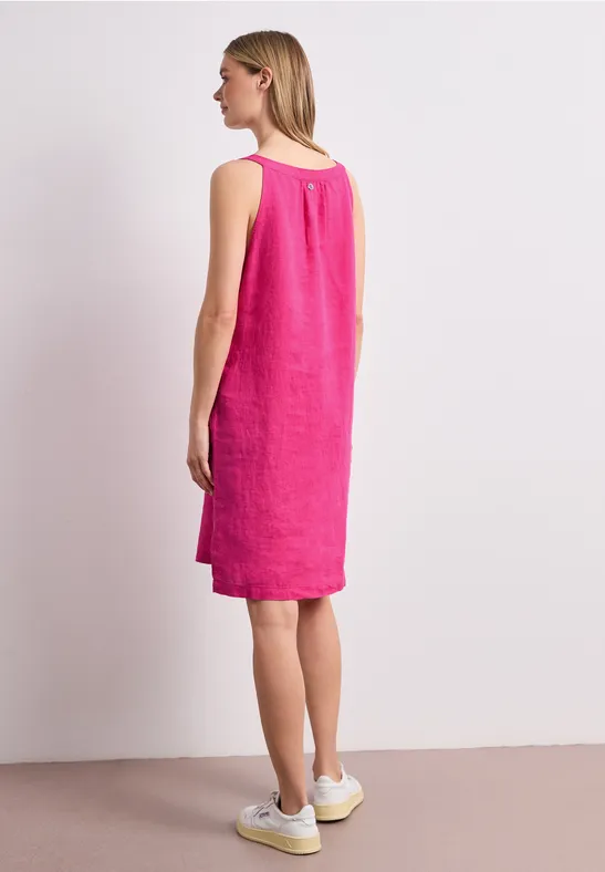 Vrouw van opzij in een knielange, mouwloze linnen jurk fuchsia roze met witte sneakers