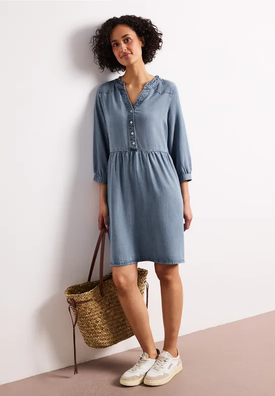 Frau trägt ein lässiges Jeanskleid mit Knopfleiste, weiße Sneaker und eine geflochtene Korbtasche
