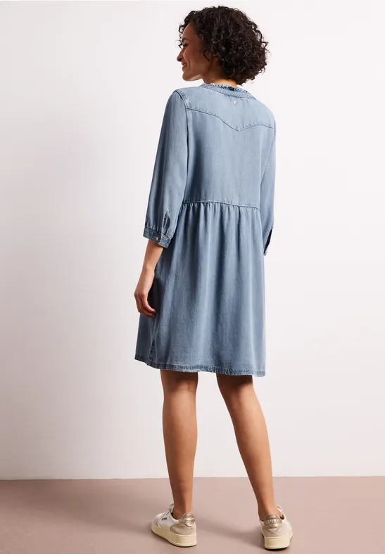 Frau von hinten in lockerem, knielangem Jeanskleid mit 3/4-Ärmeln und Sneakers vor hellem Hintergrund.