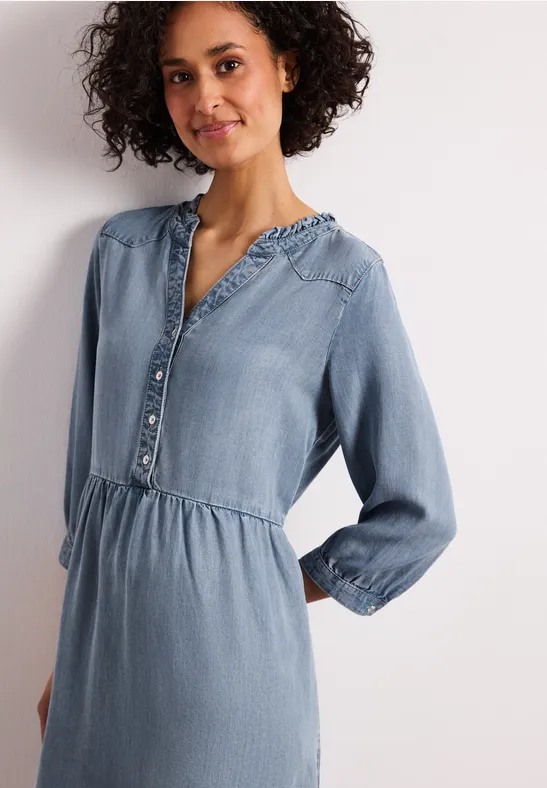 Frau in hellblauem Jeanskleid mit Knopfleiste, elastischer Taillennaht und langen Ärmeln.