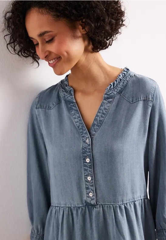 Frau in locker sitzendem hellblauen Jeanskleid mit Rüschkragen und Knopfleiste.