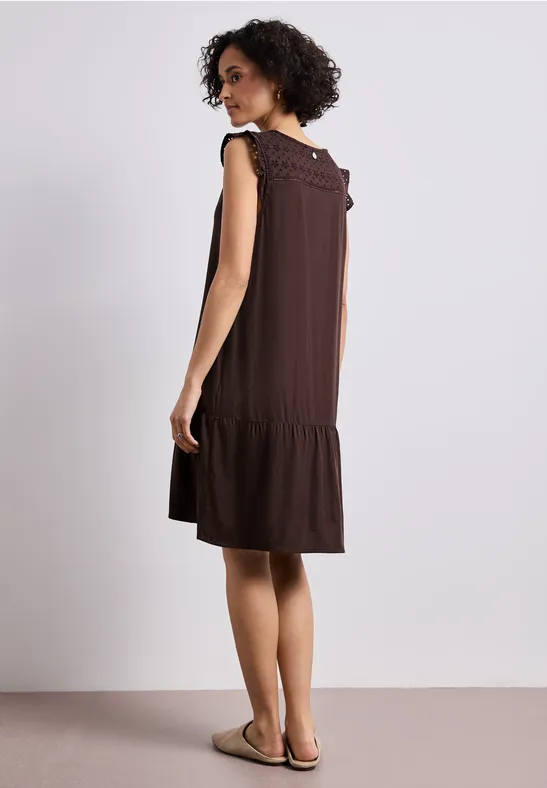 Frau in ärmellosem braunen Kleid mit Volant-Details, steht seitlich im hellen Raum, trägt bequeme Sandalen.