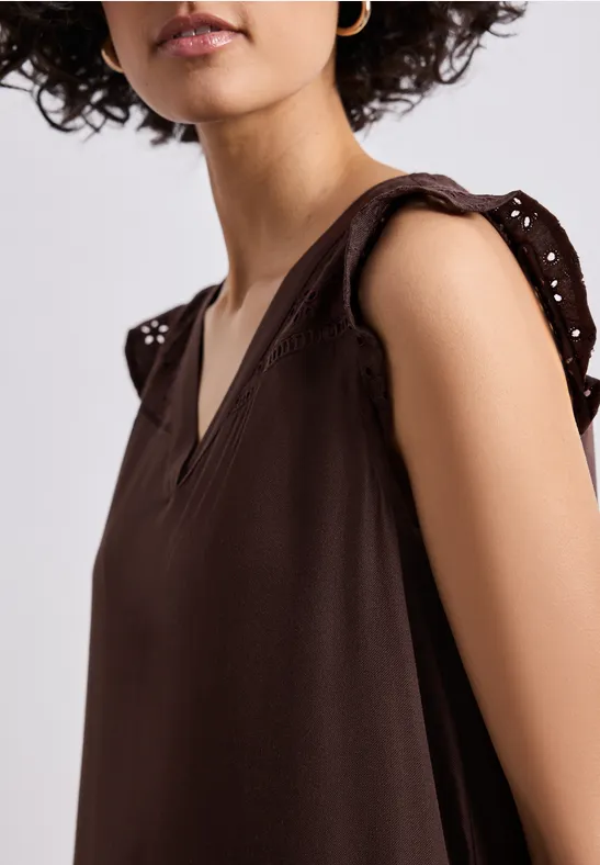 Frau in braunem, ärmellosem V-Ausschnitt-Oberteil mit dekorativen Lochstick-Ärmeln.