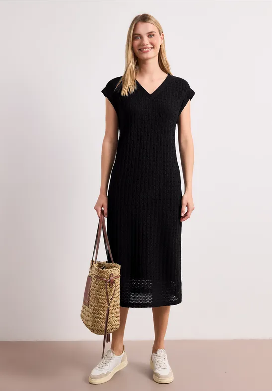 Vrouw in zwarte gebreide midadjurk met V-hals, witte sneakers en een gevlochten strooien tas.