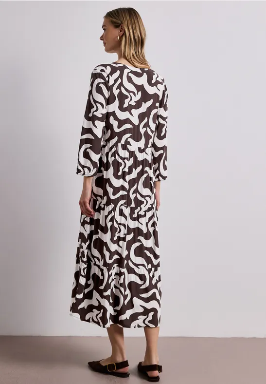 Vrouw in lange, losse jurk met bruin-wit abstract dessin voor neutrale achtergrond.
