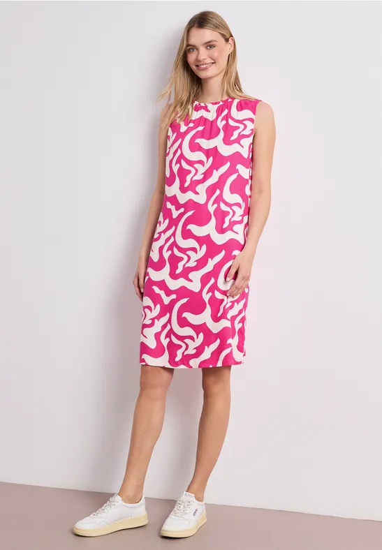 Vrouw in mouwloze midi-jurk met roze-wit dessin en witte sneakers voor een neutrale achtergrond.