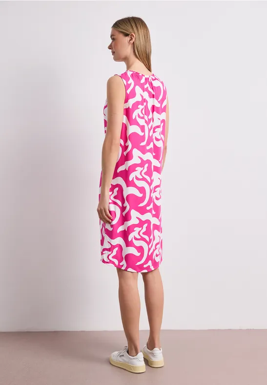 Vrouw van achteren in een knielange mouwloze jurk met roze-witte abstracte print en witte sneakers.