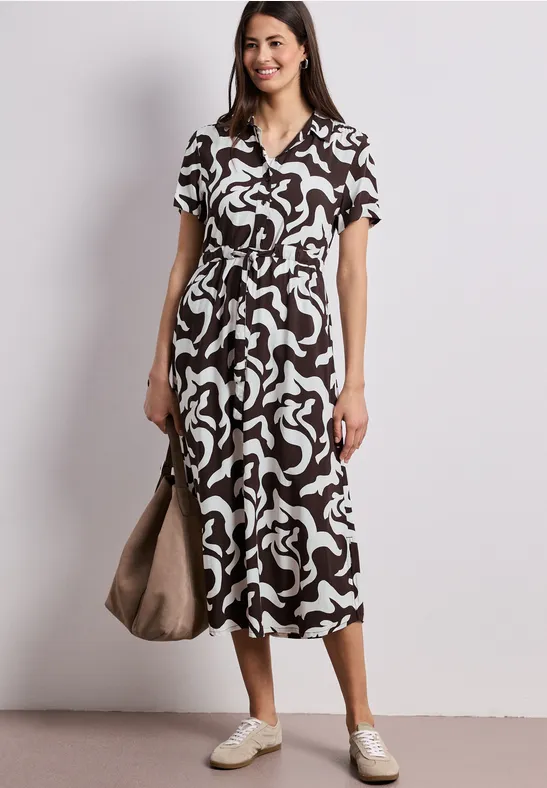 Vrouw in een midi-jurk met abstract zwart-wit patroon, V-hals, witte sneakers en tas.