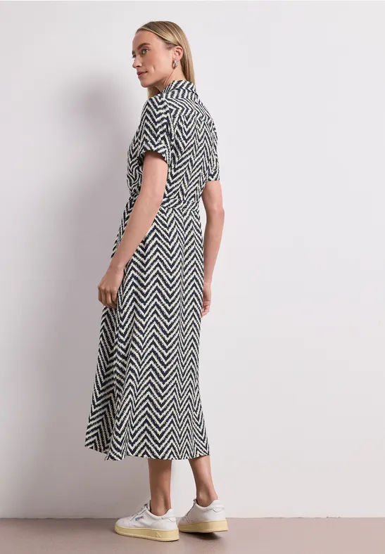 Vrouw draagt zwart-witte zigzag midi-jurk met korte mouwen en witte sneakers voor neutrale achtergrond.