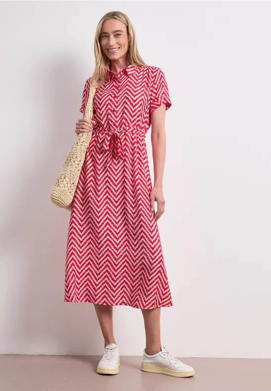 Vrouw in rood midi-jurk met zigzagpatroon, korte mouwen, rieten tas en witte sneakers tegen lichte achtergrond.