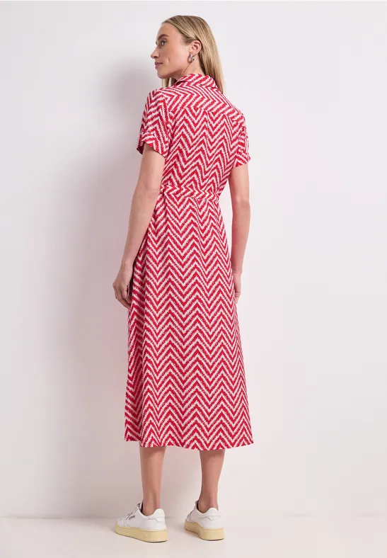 Vrouw in zijaanzicht draagt rood-witte midi-jurk met zigzagpatroon en witte sneakers voor een lichte achtergrond.