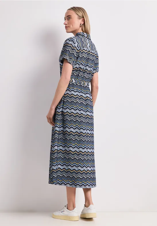 Vrouw in midijurk met blauw-wit-zwart zigzagpatroon, zijaanzicht tegen witte achtergrond.