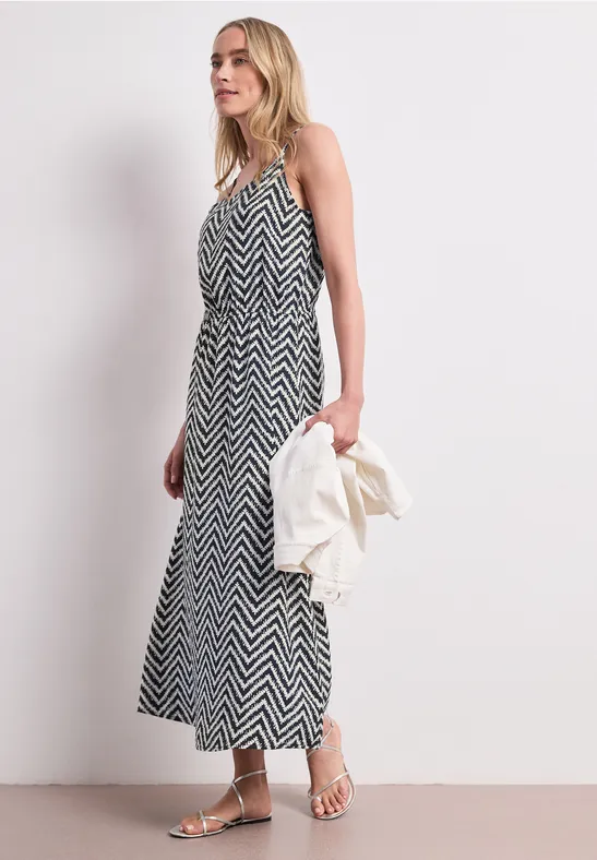 Femme en robe midi zigzag noir et blanc, tenant une veste blanche, sandales aux pieds.