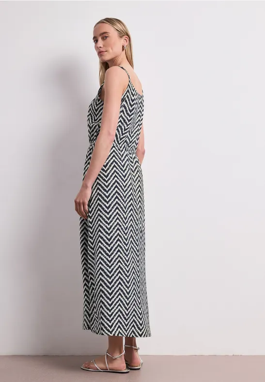 Vue de profil d'une femme portant une robe longue noire et blanche à motif zigzag et bretelles fines sur fond blanc.