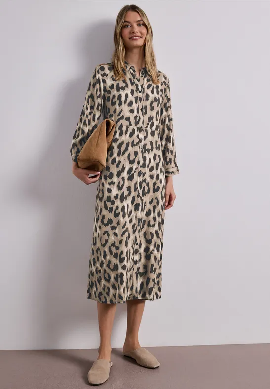 Frau in knielangem, langärmligem Leopardenmuster-Kleid mit beigefarbenen Schuhen und Clutch vor neutralem Hintergrund.