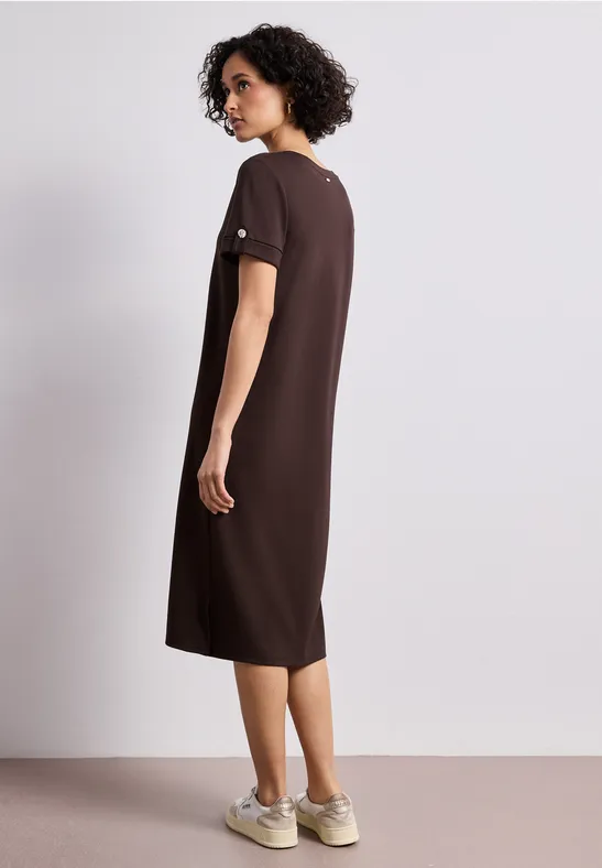 Vrouw van achteren in losvallende bruine midi-jurk en sneakers, tegen neutrale achtergrond.