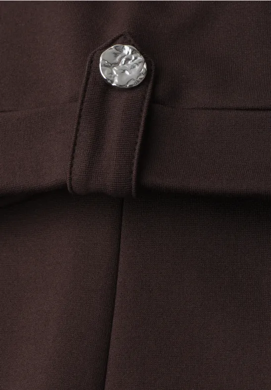 Close-up van een donkerbruin kledingstuk met een zilveren, muntvormige knop en een riemlus.