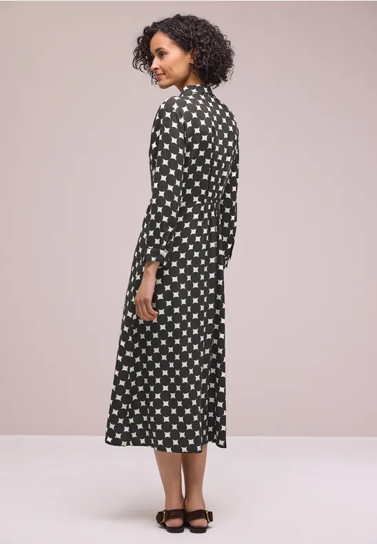 Frau in schwarzem Midikleid mit weißen Punkten, seitlich gewandt, schwarze Sandaletten