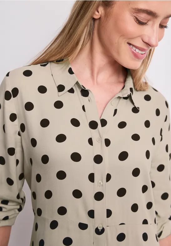 Chemisier beige à pois noirs, col et patte de boutonnage, manches retroussées de façon décontractée.