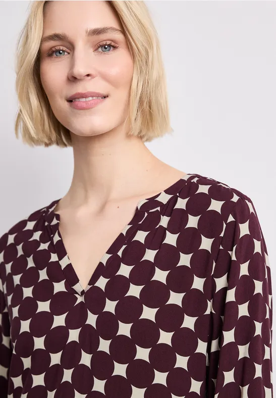 Eine Frau mit kurzem blondem Bob trägt eine bordeauxrote Bluse mit großen cremefarbenen Punkten und V-Ausschnitt.