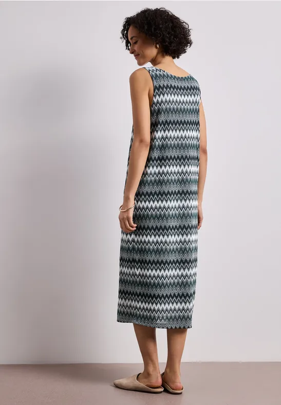 Vrouw in maxi jurk met zigzagpatroon en sandalen, staand voor een effen achtergrond.