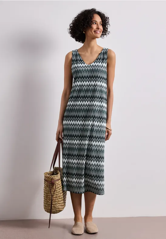 Vrouw draagt mouwloze jumpsuit met V-hals in zwart-wit-groen zigzagpatroon, strozak en beige schoenen.