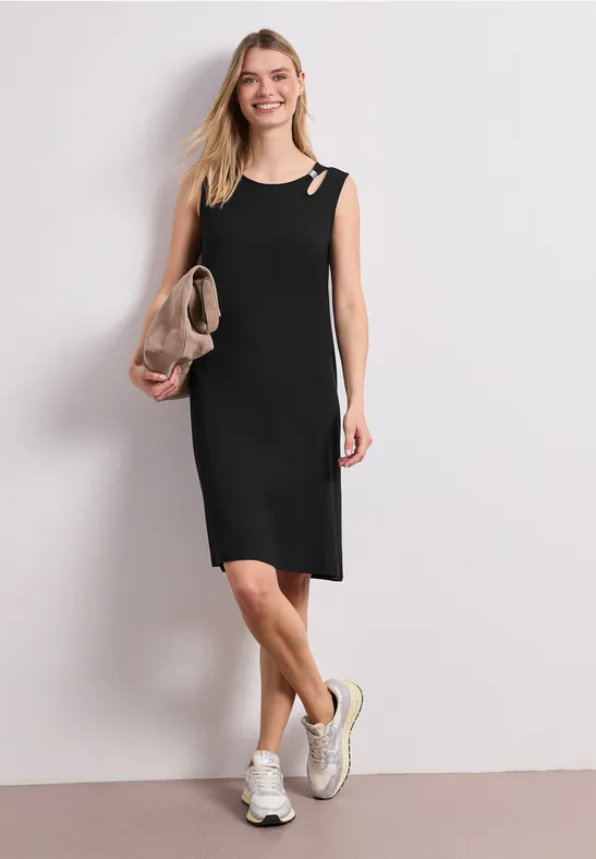 Vrouw in mouwloze zwarte jurk met clutch en sneakers voor een lichte achtergrond.