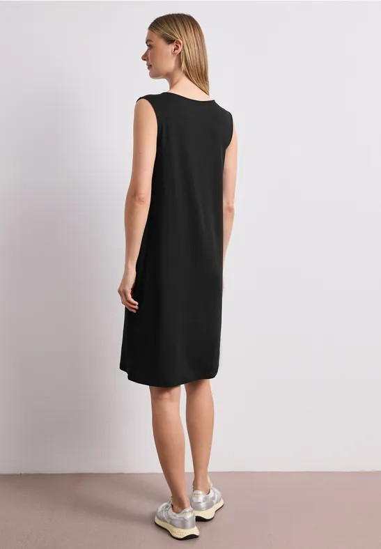 Vrouw van achter in mouwloze zwarte knielange jurk en lichtgekleurde sneakers tegen neutrale achtergrond.