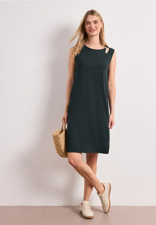 Femme en robe noire épurée sans manches à découpe épaule, sac en paille et sandales blanches.