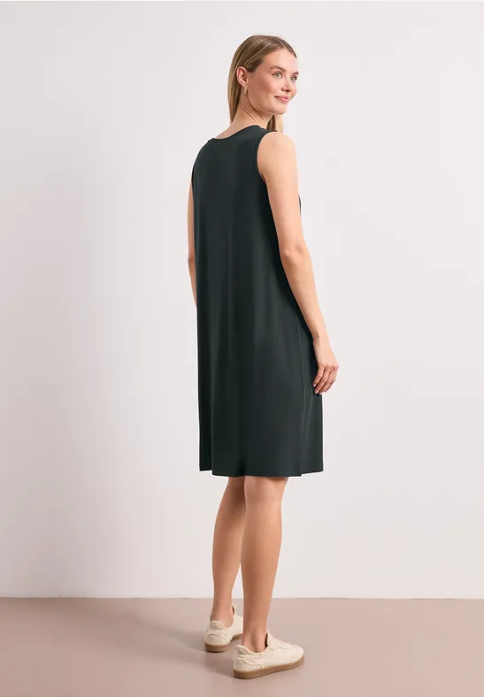 Une femme porte une robe noire sans manches jusqu'aux genoux et des baskets, vue de côté.