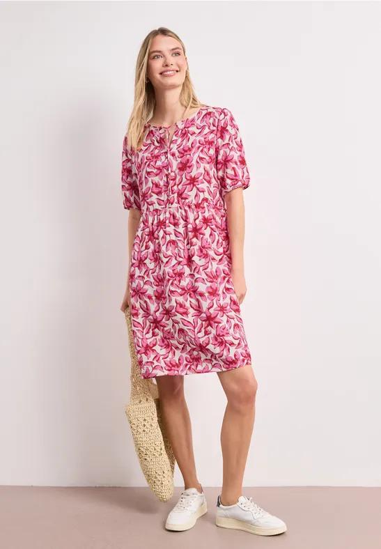Frau trägt locker sitzendes rosa-weißes Blumenkleid, weiße Sneaker, Strohtasche