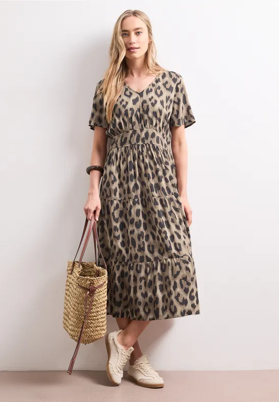 Vrouw in midi-jurk met luipaardprint, draagt een rieten tas en comfortabele beige sneakers.