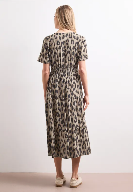 Achteraanzicht van een vrouw in een midi-jurk met luipaardprint, korte mouwen en beige schoenen voor een witte muur.