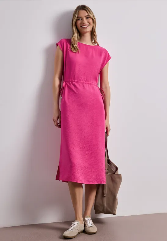 Een vrouw draagt een roze midi-jurk, witte sneakers en houdt een bruine tas vast.