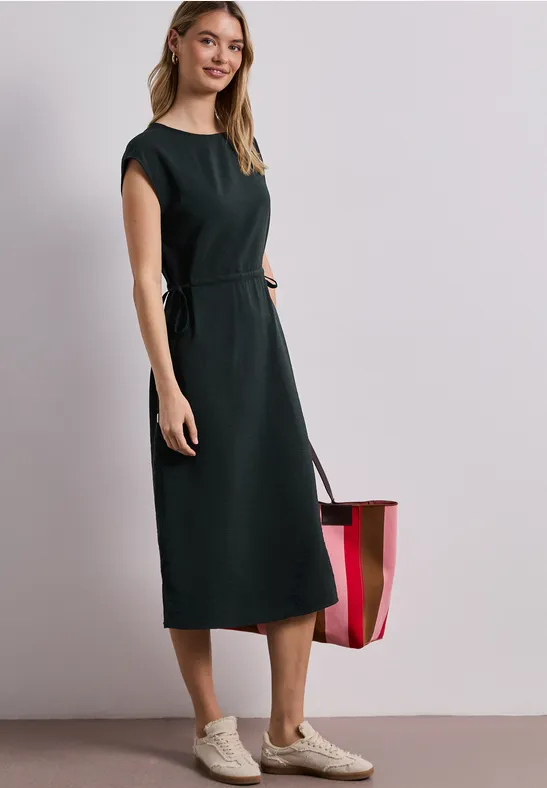 Vrouw in zwarte mouwloze midi-jurk met ceintuur, crèmekleurige sneakers en gestreepte draagtas.