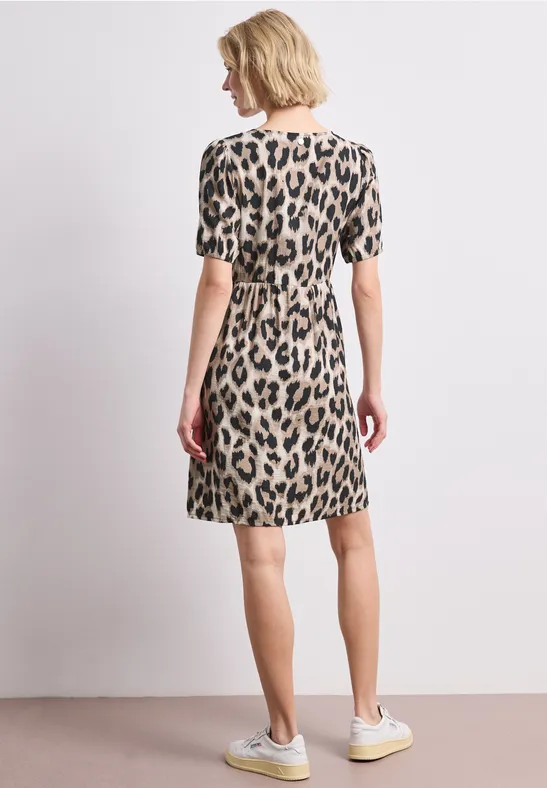 Vrouw van achteren in korte luipaardprint jurk met lichte sneakers tegen een neutrale achtergrond.