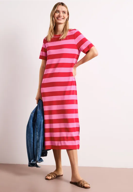 Frau in Midi-T-Shirt-Kleid mit rosa-roten Streifen und flachen Sandalen, in der Hand eine Jeansjacke