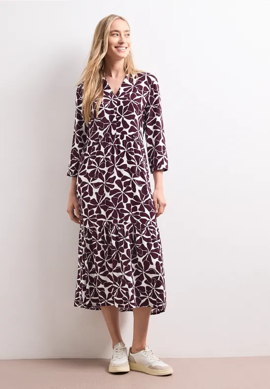 Blonde Frau trägt ein violett-weiß gemustertes Midikleid und weiße Sneaker vor weißem Hintergrund.