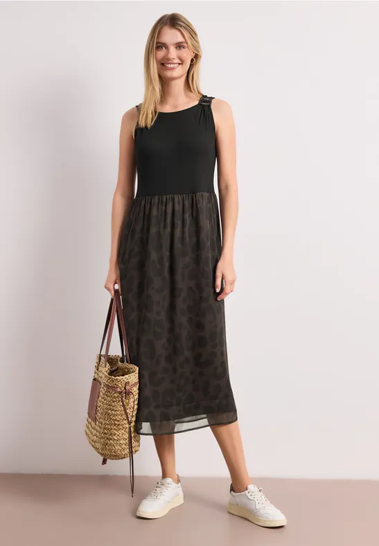 Een vrouw in een mouwloos zwart topje, een midi rok met print, witte sneakers en een gevlochten tas
