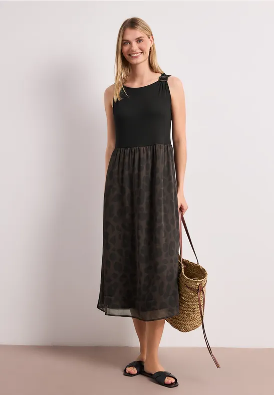 Vrouw in zwarte mouwloze top, lange rok met print en strooien tas, voor een lichte achtergrond.