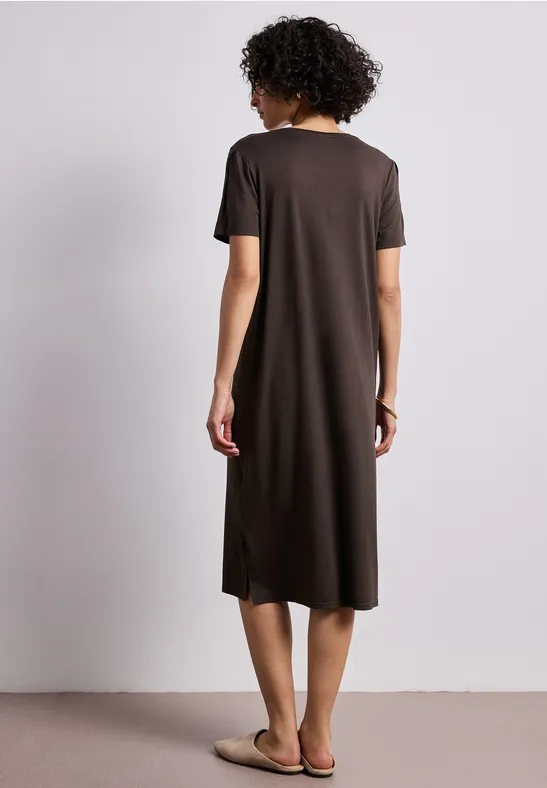 Vrouw met kort krullend haar van achteren in een bruine midi-jurk en sandalen tegen een neutrale achtergrond.