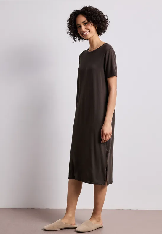 Een vrouw draagt een eenvoudig donkerbruin midi-jurk met korte mouwen en comfortabele pantoffels voor een neutrale achtergrond.