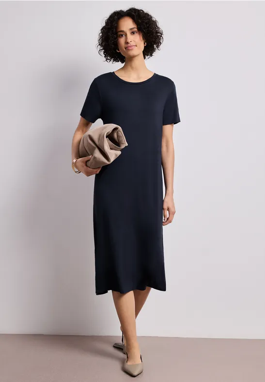 Femme portant une robe T-shirt bleu marine jusqu’aux genoux, tenant un manteau beige, avec des chaussures plates.