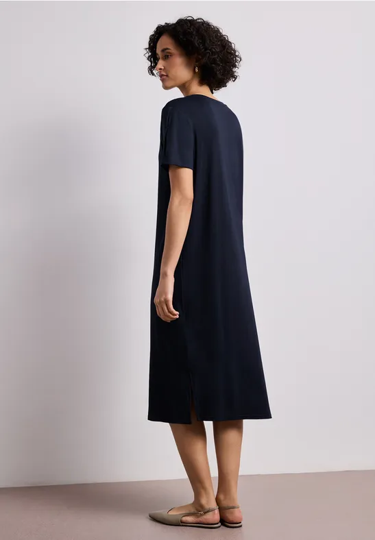 Femme de dos, porte une robe midi bleue marine à manches courtes, fente au dos, sandales plates.